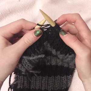 May include: Close-up of a person knitting with two wooden needles. The knitting project is a striped black and gray fabric.