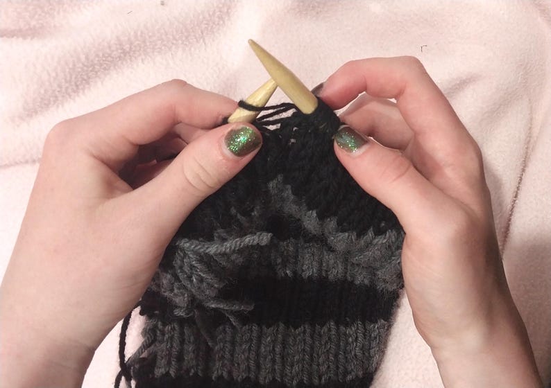 May include: Close-up of a person knitting with two wooden needles. The knitting project is a striped black and gray fabric.