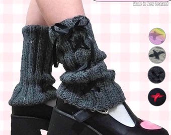May include: Gray knitted leg warmers with black ribbon ties and a bow. The leg warmers are worn over black platform shoes with a white toe and a pink heart on the side. The text "100% WOOL Made in New Zealand" is displayed in the upper right corner of the image.