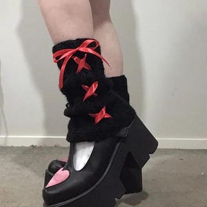 May include: Black platform shoes with a pink heart detail on the toe. The shoes have a white trim and are worn with black fuzzy leg warmers with red ribbon bows.