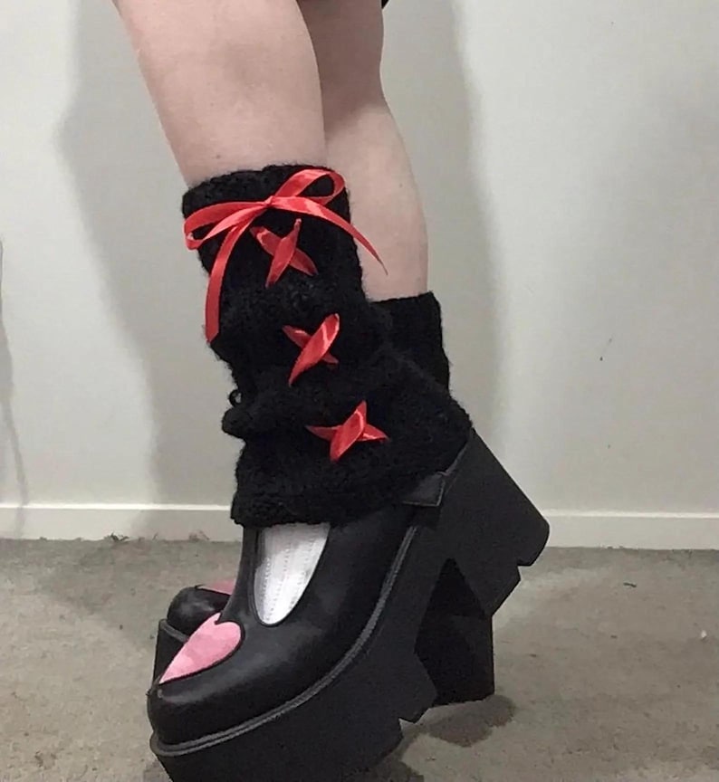 May include: Black platform shoes with a pink heart detail on the toe. The shoes have a white trim and are worn with black fuzzy leg warmers with red ribbon bows.
