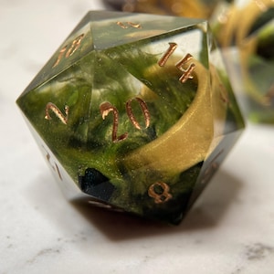 May include: A green and gold 20-sided dice with a gold ring embedded inside. The dice has numbers 2, 8, 14, and 20 visible.