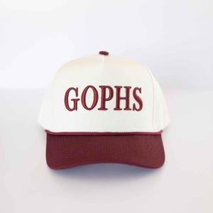 May include: A white and maroon baseball cap with the word "GOPHS" embroidered in maroon. The cap has a maroon brim and a maroon rope detail along the bottom edge of the white crown.