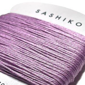 May include: A spool of lavender-colored sashiko thread, 100% cotton, with a label that reads "SASHIKO" and "30m".