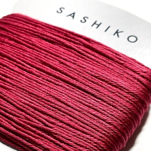 Puede incluir: Una bobina de hilo de sashiko rojo carmesí de 30 metros. El hilo está enrollado alrededor de una tarjeta blanca con la palabra "SASHIKO" impresa en negro.