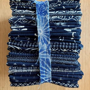 May include: A stack of folded indigo blue fabric squares with white patterned designs. A light blue ribbon with a tie-dye pattern wraps around the stack. The fabric is arranged on a light brown wooden surface.