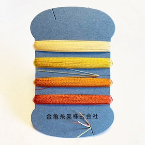 May include: A close-up of a spool of thread with four different colors: cream, yellow, orange, and red. The spool is labeled "金銀糸業株式会社" in Japanese.