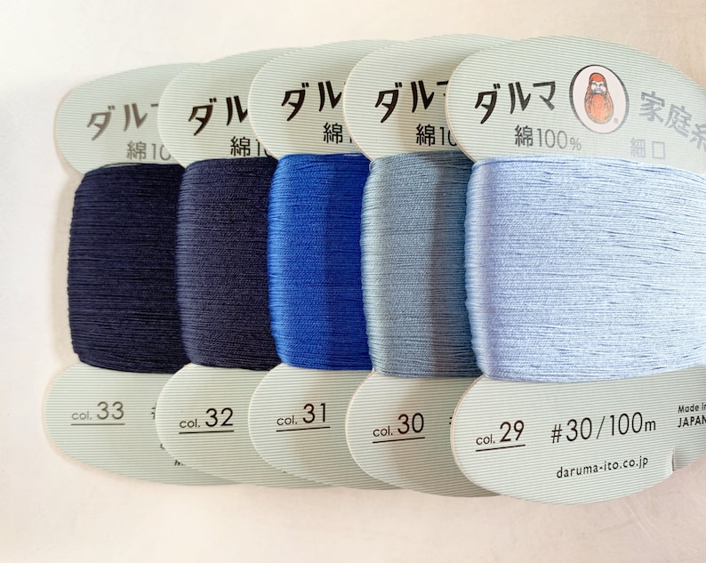 Op de afbeelding: Vijf spoelen blauwe katoenen draad, gelabeld met het Japanse woord "ダルマ" (Daruma) en de tekst "綿100%" (katoen 100%). De spoelen zijn genummerd 33, 32, 31, 30 en 29. De laatste spoel heeft ook de tekst "#30/100m" en "daruma-ito.co.jp".