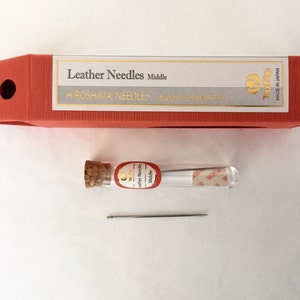 May include: A set of leather needles, packaged in a red box with the text "Leather Needles Middle" and "Hiroshima Needles". A small glass vial with a cork stopper contains several needles. A single needle lies below the vial.