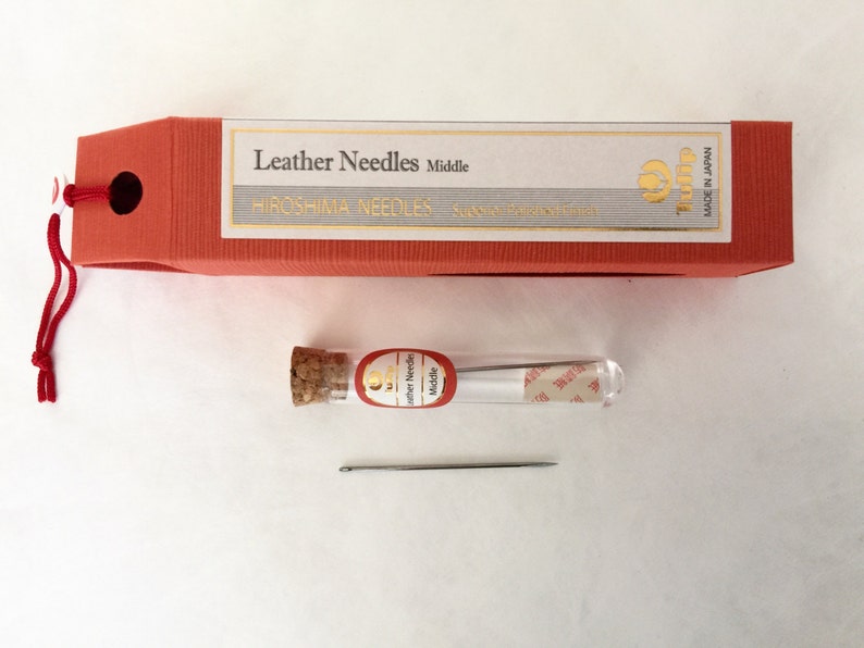 May include: A set of leather needles, packaged in a red box with the text "Leather Needles Middle" and "Hiroshima Needles". A small glass vial with a cork stopper contains several needles. A single needle lies below the vial.