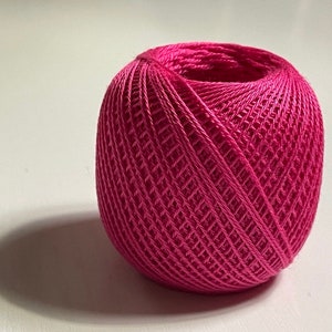 May include: A ball of vibrant fuchsia-colored crochet thread. The thread is tightly wound, creating a textured, spherical shape. The thread's intricate woven pattern is visible, and the ball casts a shadow on the white surface.