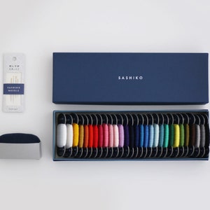 May include: A blue box with the word "SASHIKO" on the lid, containing a set of 24 spools of colorful embroidery floss. The spools are arranged in a row, with each spool containing a different color of thread. The box also includes a small package of embroidery needles.