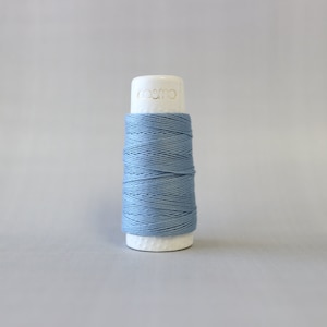 May include: A spool of light blue thread, tightly wound around a white cylindrical core. The top of the core is labeled "cosmo" in gold lettering. The thread is a solid, muted blue color, suitable for various sewing and embroidery projects.