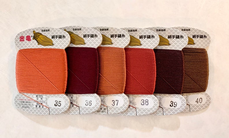 May include: A collection of six spools of thread in various shades of brown, ranging from light orange to dark brown. Each spool is labeled with a number from 35 to 40. The packaging includes Japanese text and a gold turtle logo.