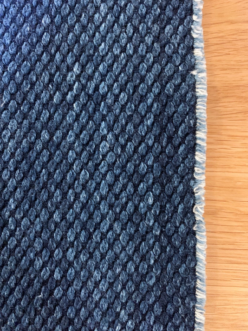 May include: Close-up of a dark blue fabric with a textured pattern of small, raised circles. The fabric has a white fringe along the edge.