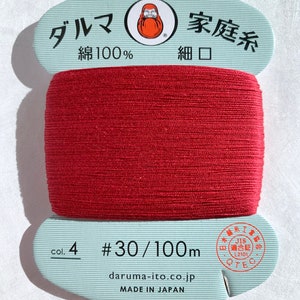 May include: A spool of red cotton thread with Japanese text and a Daruma doll logo. The label reads "col. 4 #30/100m" and "MADE IN JAPAN". The thread is tightly wound, showing the texture of the fibers.
