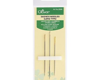 Puede incluir: Un paquete de agujas Sashiko Clover (tipo largo). El paquete tiene un diseño verde y blanco con el logotipo de Clover y texto en varios idiomas. Se ven tres agujas, sujetas por una pieza de espuma blanca.