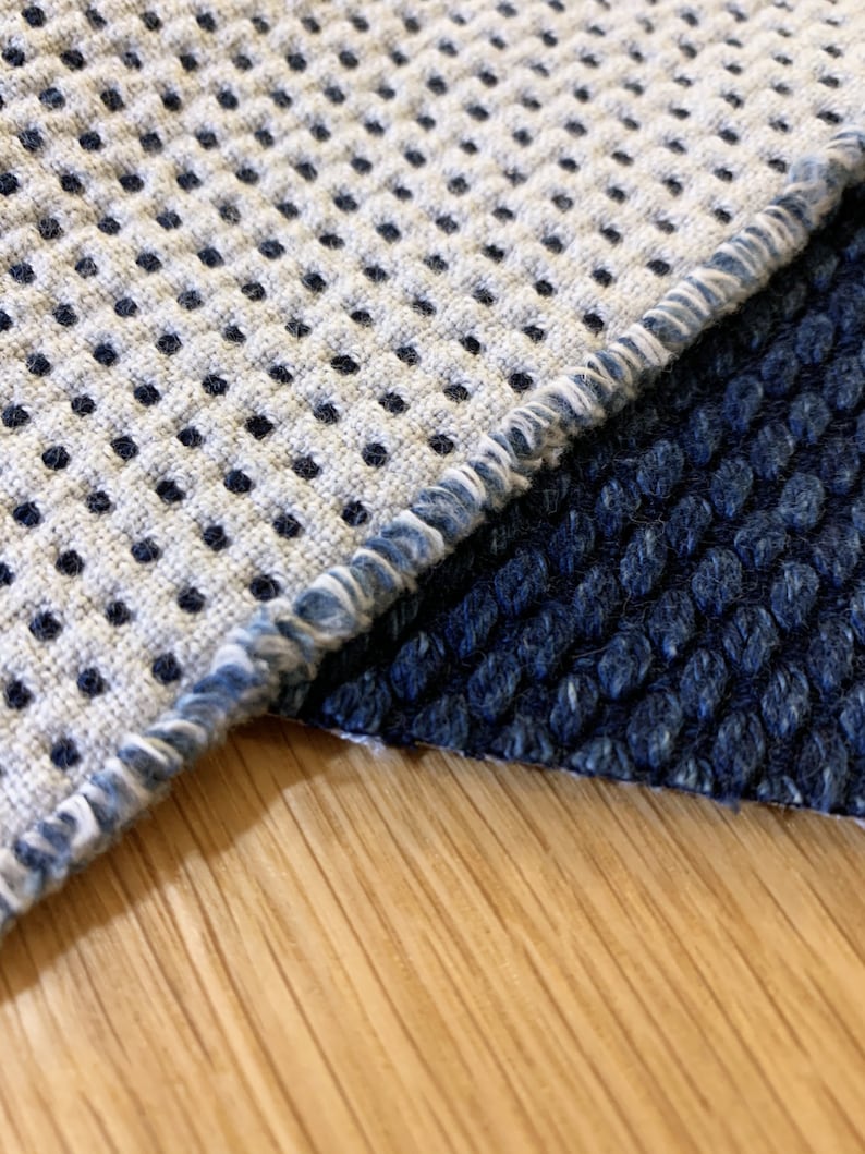 May include: Close-up of two fabric swatches with different textures. The top swatch is a light gray with a black polka dot pattern and a frayed edge. The bottom swatch is a dark blue with a textured surface. The swatches are on a light wood surface.