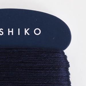 May include: Close-up of a spool of dark blue Sashiko thread. The thread is tightly wound, and the label above reads "SASHIKO" in white capital letters against a dark blue background. The background is white.