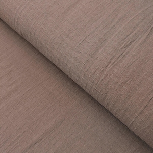 May include: Close-up of a folded, textured fabric in a muted brown color. The fabric has a subtle grid pattern and a soft, natural appearance, suggesting it is made of linen or a similar material. The folds create shadows and depth.