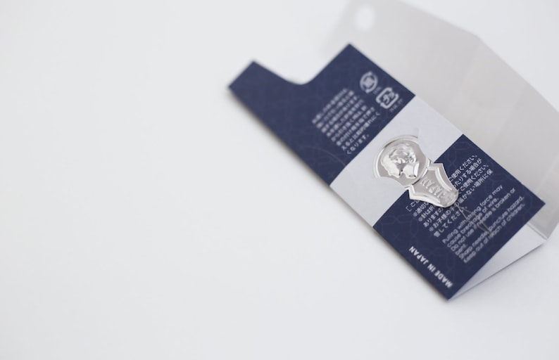 May include: A silver key-shaped bookmark with the words "MY SHELF" engraved on it. The bookmark is attached to a blue and white card with Japanese text and the words "MADE IN JAPAN".