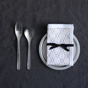 Puede incluir: Una servilleta de lino blanco con un patr&oacute;n geom&eacute;trico en hilo negro y dorado est&aacute; atada con una cinta negra y colocada sobre un plato de plata. Un tenedor y una cuchara se colocan a la izquierda del plato.