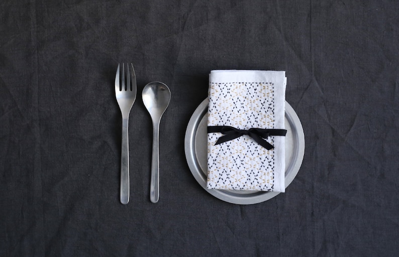 Puede incluir: Una servilleta de lino blanco con un patr&oacute;n geom&eacute;trico en hilo negro y dorado est&aacute; atada con una cinta negra y colocada sobre un plato de plata. Un tenedor y una cuchara se colocan a la izquierda del plato.