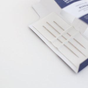 May include: A close-up of a white box with a blue trim, containing a set of five silver sewing needles.