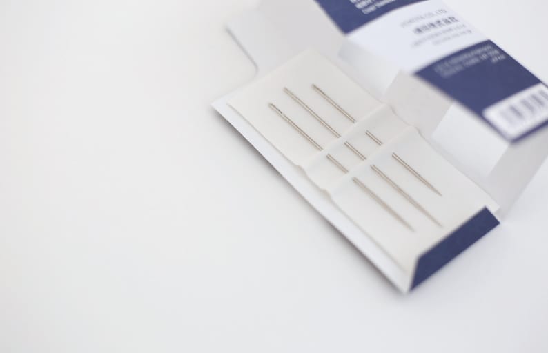 May include: A close-up of a white box with a blue trim, containing a set of five silver sewing needles.
