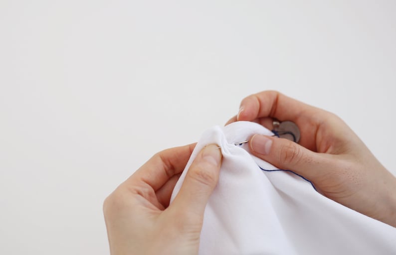 May include: A person's hands are shown sewing a white fabric with a needle and blue thread. The needle is threaded through the fabric and the thread is pulled through. The person is using their fingers to hold the fabric in place.