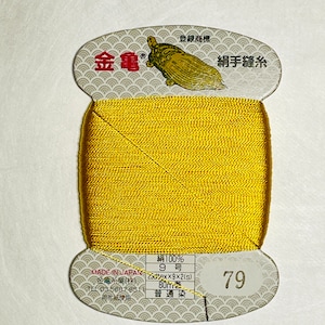 May include: A spool of gold-coloured thread with a label that reads "絹手縫糸" (silk hand sewing thread) and "金亀" (Kin-Kame) in Japanese. The label also includes the text "絹100%" (100% silk), "9号" (9 gauge), "23dtex×9×2(s)", "80m巻" (80m roll), "普通染" (ordinary dyeing), and "79".