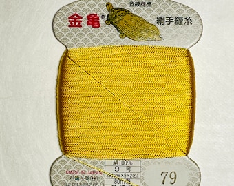 May include: A spool of gold-coloured thread with a label that reads "絹手縫糸" (silk hand sewing thread) and "金亀" (Kin-Kame) in Japanese. The label also includes the text "絹100%" (100% silk), "9号" (9 gauge), "23dtex×9×2(s)", "80m巻" (80m roll), "普通染" (ordinary dyeing), and "79".