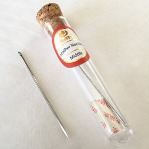 May include: A clear glass vial with a cork stopper containing several silver leather needles. One needle is outside the vial. The vial has a red and gold label that reads "Leather Needles Middle".