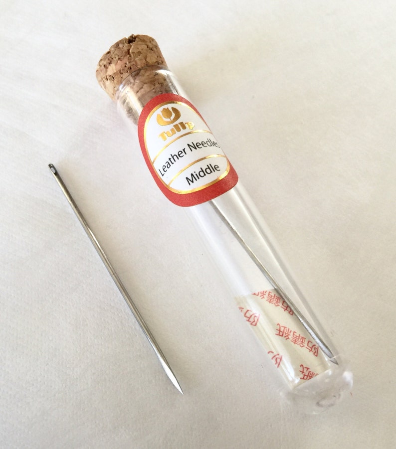 May include: A clear glass vial with a cork stopper containing several silver leather needles. One needle is outside the vial. The vial has a red and gold label that reads "Leather Needles Middle".