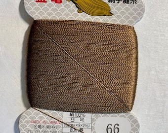 Puede incluir: Una bobina de hilo de seda marrón con una etiqueta que dice "絹手縫糸" (hilo de coser a mano de seda) y "金亀" (Kinkame) en japonés. La etiqueta también incluye el texto "Made in Japan", "絹100%" (100% seda), "9号" (9 calibre), "23dtex×9×2(s)", "80m巻" (bobina de 80 m), "普通染" (teñido liso) y "66".