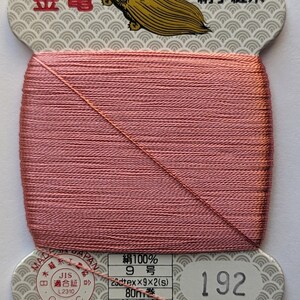 May include: A spool of pink thread, labelled with the number 192. The label features Japanese text and a gold turtle logo. The thread is tightly wound and appears to be silk. The spool is made in Japan.