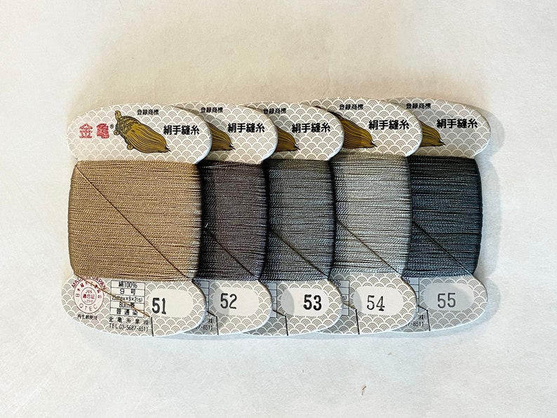 May include: Five spools of thread in varying shades of brown and gray, labeled 51 through 55. Each spool is neatly wound and attached to a decorative label with Japanese text and a gold emblem. The thread is likely for sewing or embroidery.