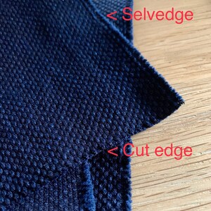 以下が含まれることがあります： 表面がテクスチャ加工された濃い青色の生地のクローズアップ。生地は耳とカットエッジを持っています。