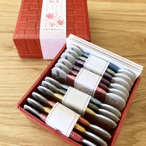 May include: A red box with a woven pattern and a label that says "秋色" in Japanese characters. The box contains a set of eight spools of embroidery floss, each wrapped in a white paper with a label. The floss is in various colors, including gold, silver, and shades of red, green, and blue.