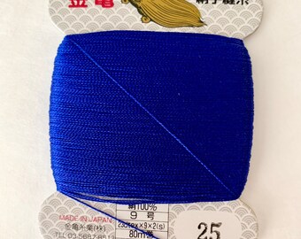 May include: A spool of bright blue sewing thread, with Japanese text and the number 25 on the label. The label features a gold turtle illustration and the text "MADE IN JAPAN". The thread is tightly wound on a white cardboard spool.