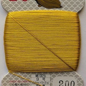 May include: A spool of gold-colored thread with a label that reads "絹100% 9号 26dtex×9×2(s) 80m巻 普通染 絹手縫糸 200" and "MADE IN JAPAN" and "金亀糸業(株) 03-5687-8511 再生紙使用".