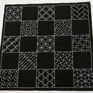 May include: A black square textile with a grid pattern of white embroidered designs. The designs vary in each square, including floral, geometric, and wave patterns. The edges are neatly finished with a white border.