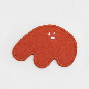 May include: A red felt patch in the shape of a blob with a white embroidered face. The patch has a simple design with two white dots for eyes and a small white oval for a mouth.