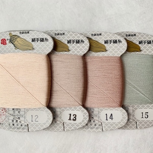 May include: Four spools of silk thread in shades of beige, taupe, rose, and sage green. Each spool is labeled with a number and Japanese text. The thread is tightly wound on a rectangular card with a decorative top.