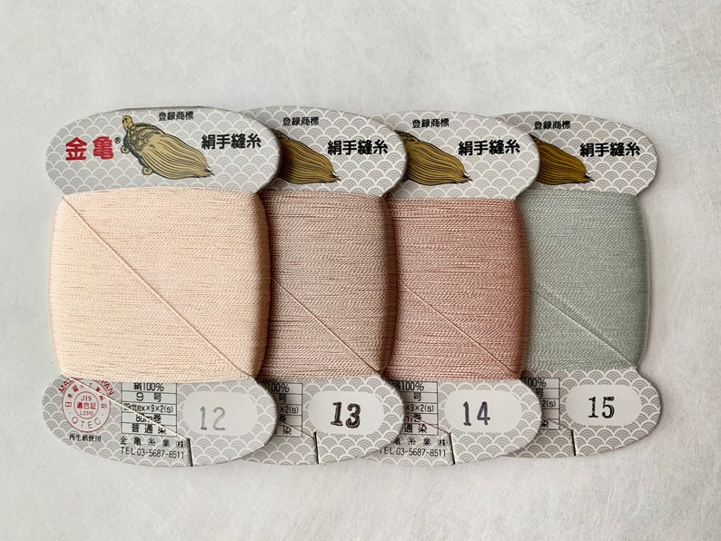 May include: Four spools of silk thread in shades of beige, taupe, rose, and sage green. Each spool is labeled with a number and Japanese text. The thread is tightly wound on a rectangular card with a decorative top.