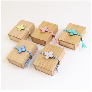May include: Five small wooden boxes with different colored fabric flowers tied on top. The boxes are all made of light brown wood and have a natural finish.
