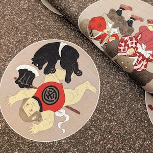 May include: Japanese-style fabric with a repeating pattern of figures and a bear. The figures are depicted in a traditional style, with bold colors and intricate details. The fabric has a textured, speckled background in shades of brown.