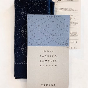 Puede incluir: Un kit de bordado sashiko azul y blanco con instrucciones. El kit incluye un panel de tela preimpreso con un patr&oacute;n de diamante triple, una aguja e hilo. El embalaje presenta el texto japon&eacute;s "DARUMA SASHIKO SAMPLER 刺し子ふきん 三重菱つなぎ SANJ&Uuml;BISHI-TSUNAGI Triple Diamond Pattern".