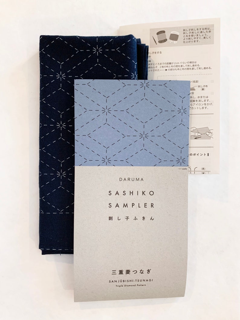 Puede incluir: Un kit de bordado sashiko azul y blanco con instrucciones. El kit incluye un panel de tela preimpreso con un patr&oacute;n de diamante triple, una aguja e hilo. El embalaje presenta el texto japon&eacute;s "DARUMA SASHIKO SAMPLER 刺し子ふきん 三重菱つなぎ SANJ&Uuml;BISHI-TSUNAGI Triple Diamond Pattern".