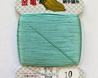 May include: A spool of light green silk thread with a white label that reads "絹100% 9号 23dtex×9×2(s) 80m巻 普通染" and "金亀糸業(株) TEL 03-5687-8511 再生紙使用" and "10". The label also features a turtle logo and the text "登録商標 金亀 絹手縫糸".